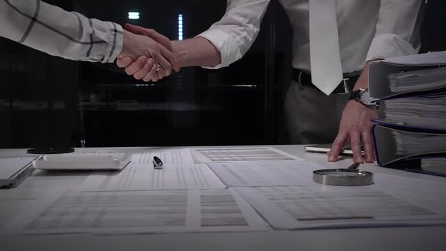Business professionals shake hands over a cluttered desk of contracts, calculator and magnifying glass, sealing a trusted partnership after detailed financial review and late night work