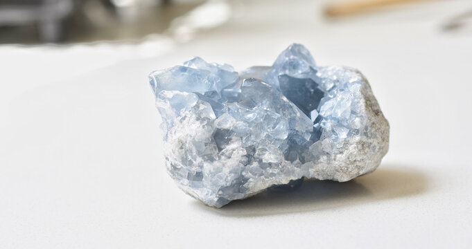 High-quality image of a natural celestite geode crystal resting on a clean white table. This blue celestine mineral features delicate sparkling formations and soft pastel tones