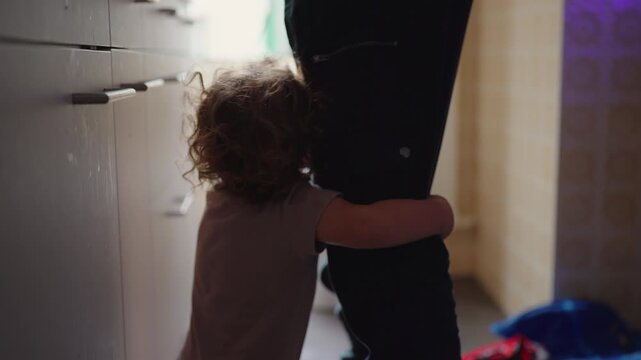 Toddler crying and clinging to caregiver leg in kitchen seeking attention and wanting to be picked up emotional childhood moment during everyday home routine