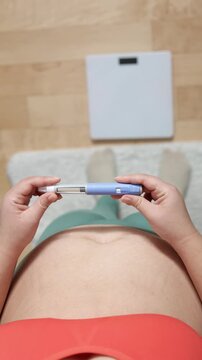 A vertical close up of an young woman giving an injection to her belly
A young woman giving an injection of semaglutide to her belly on floor and scales background