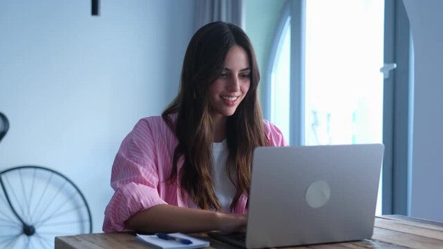 A cheerful young woman diligently works on her laptop at home, occasionally pausing to jot down notes. Her focused expression suggests she is engaged in a productive task, whether for business 