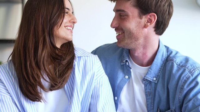 A lovely young couple relaxes at home, sharing tender glances and warm smiles. They enjoy a peaceful moment, showcasing their strong bond and mutual affection and happiness.