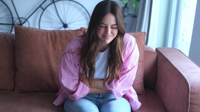 A young woman experiences acute stomach pain and discomfort while relaxing on her living room sofa. She holds her abdomen, trying to find relief from the painful symptoms of indigestion 