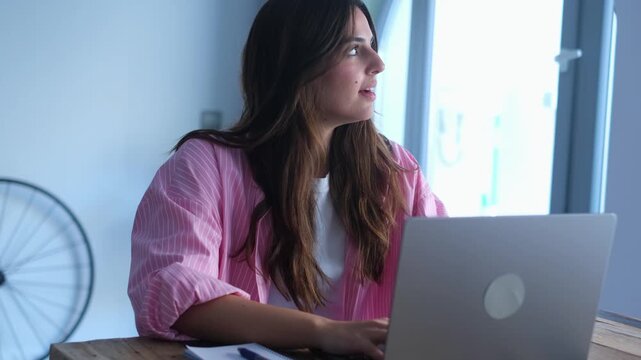 A focused young woman works remotely on her laptop from the comfort of her home. She momentarily pauses from typing to look out the window, contemplating new ideas and finding inspiration 