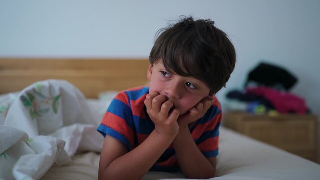 Boy lying on bed resting head on hands looking away thoughtful childhood moment of quiet reflection and calm solitude inside home