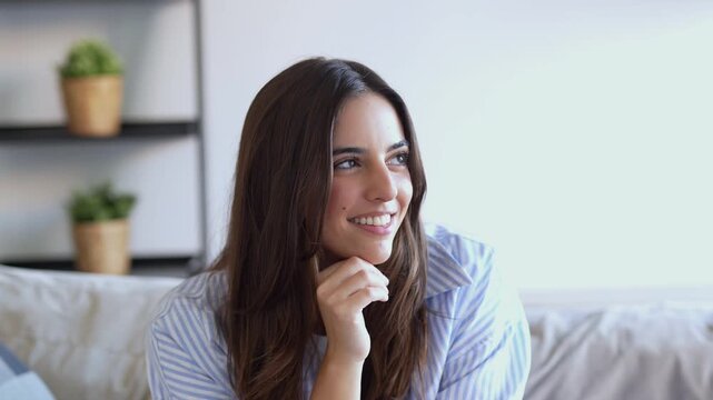 A beautiful young woman with a radiant smile expresses joy and contentment while relaxing comfortably on her sofa at home. She looks around with a cheerful expression, radiating happiness