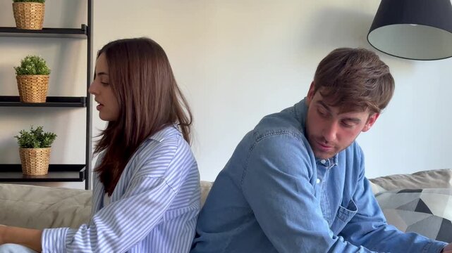 A young couple experiencing conflict sits back-to-back on a sofa. They eventually turn to face each other, engaging in a tense argument. The scene captures the breakdown of communication and relations