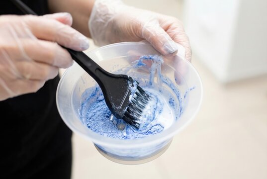 Gloved hands holding a plastic bowl and using a black brush to mix blue hair bleaching powder with developer liquid