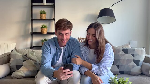 A happy young couple sits on a comfortable sofa at home, sharing a moment while looking at a smartphone. They laugh and smile, enjoying the content together.