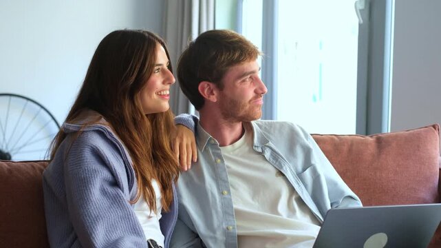A happy young couple relaxes on a sofa at home, enjoying shared moments while using a laptop. They smile and connect, finding joy in each other's company and the content they view together.