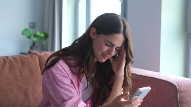 A young woman on a sofa receives upsetting news on her mobile phone, reacting with profound sadness and distress. Her emotional expression conveys disappointment and frustration.