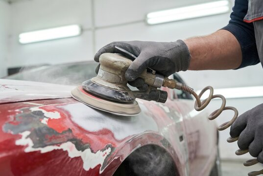 Mechanic wearing gloves using a pneumatic sanding machine to prep the red bodywork of a car