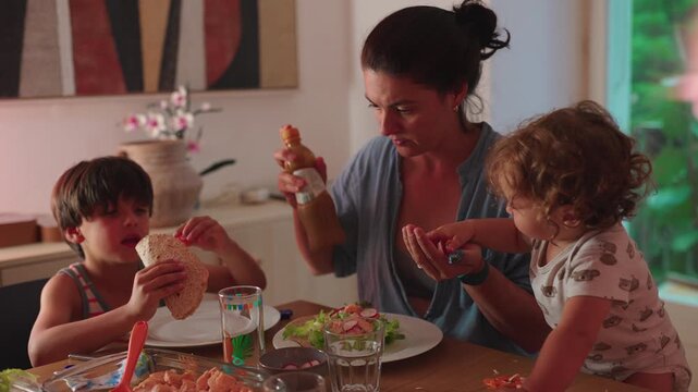 Mother sitting at table preparing salad while children eat and reach for food everyday family meal moment showing multitasking parenting and shared domestic routine