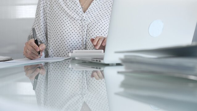 Unknown businesswoman using a calculator to crunch numbers and jotting down meticulous notes at a sleek glass desk in a contemporary office, closeup view. Taxes, audit in business
