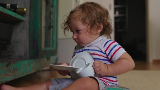 Toddler sitting on floor holding cup and plate fragile household objects creating potential risk while exploring kitchen items during everyday childhood curiosity at home