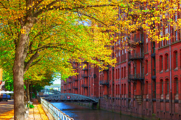 Obraz premium Speicherstadt in Hamburg features iconic red brick warehouses and canals lined with autumn trees. Historic district, UNESCO World Heritage site, showcases stunning Neo-Gothic architecture and bridges