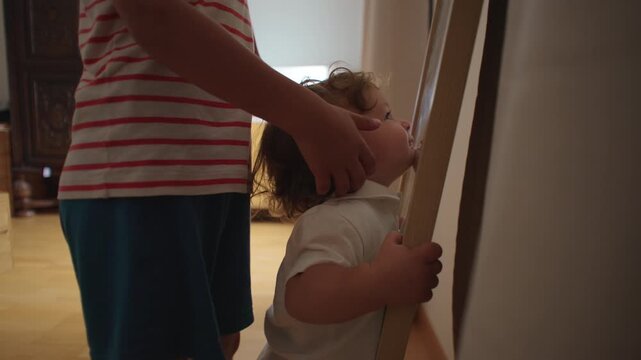 Toddler standing at mirror with sibling hand on back exploring reflection gentle childhood moment of support curiosity and early self awareness inside home