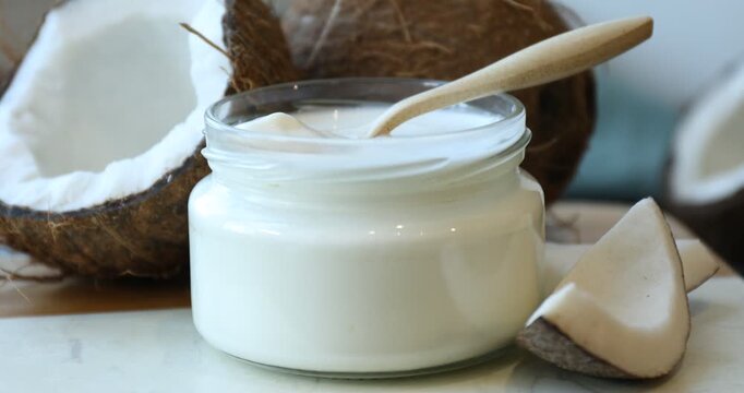 Tasty coconut yogurt in jar, spoon and fruits turning on board against blurred background, closeup