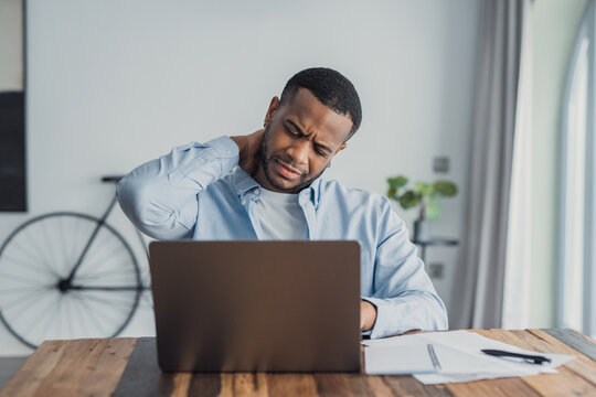 Young Black man rubs his neck while working on a laptop in a bright home office, showing frustration and physical discomfort from prolonged computer use and poor posture.