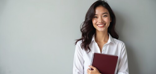 Obraz premium Smiling asian woman holds red notebook. Confident young pro with wavy dark hair wears white shirt. She looks directly at camera with friendly happy expression, ready for work.