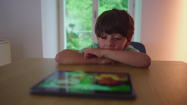 Young child resting head on arms at table while looking toward tablet, capturing boredom, passive attention, and quiet modern childhood moment in everyday home environment