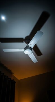 Low angle view of a white ceiling fan rotating slowly in a dark bedroom at night, casting long shadows under soft moonlight and dim glow, minimal, electric, motion blur