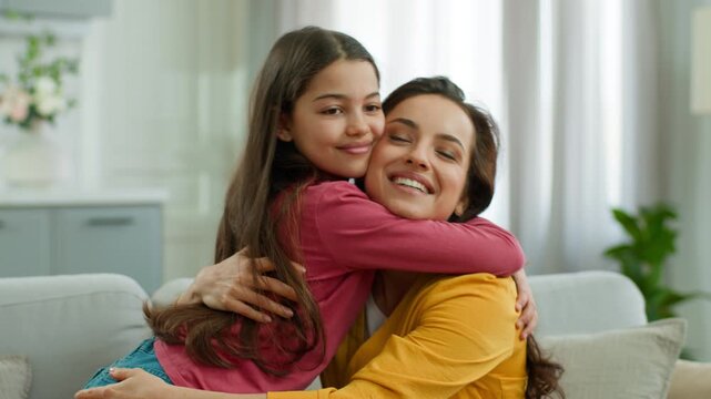 Happy Caucasian family loving little girl daughter hugging mother sister smiling affectionate woman female parenting cuddling embracing child kid laughing sitting indoor on couch at home relationship