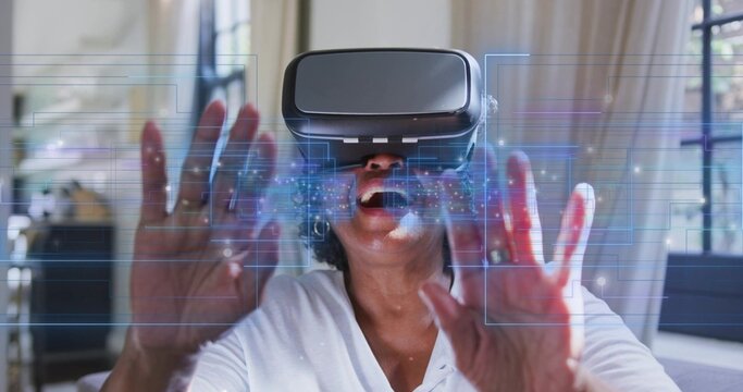 Reaching senior woman wearing white top touching holographic interface in living room, VR headset