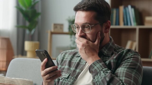 Portrait scared Caucasian man looking at mobile phone shocked reaction bad news adult shock amazed guy close mouth with hand problem online indoors male in apartment holding using smartphone at home
