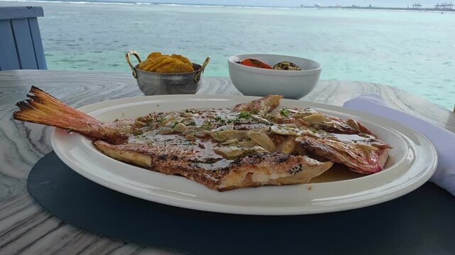 Caribbean style fish with plantains and grilled vegetables served on a  table in front of the beach.