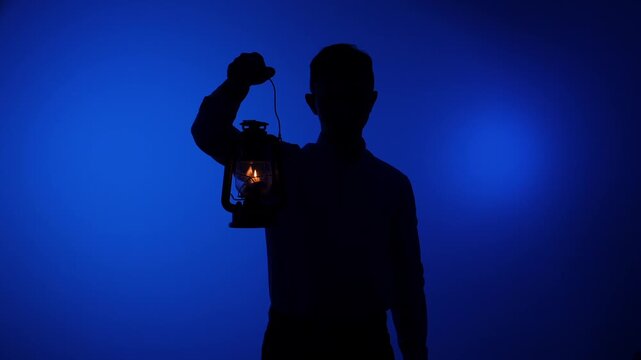 Man silhouette standing in front of a blue background, holding up a vintage oil lamp with a bright, flickering flame, symbolizing mystery, guidance, exploration, and finding a way through unknown