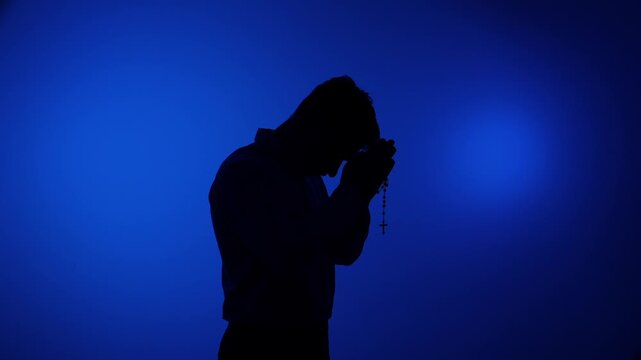 Man silhouetted kneeling in prayer, holding rosary beads against a minimalist blue background, finding solace, humility and hopeful devotion in quiet contemplation