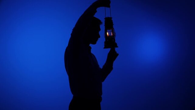 Man's silhouette standing in darkness, holding a glowing lantern and a wooden christian cross, representing faith, hope, and spirituality in search of guidance