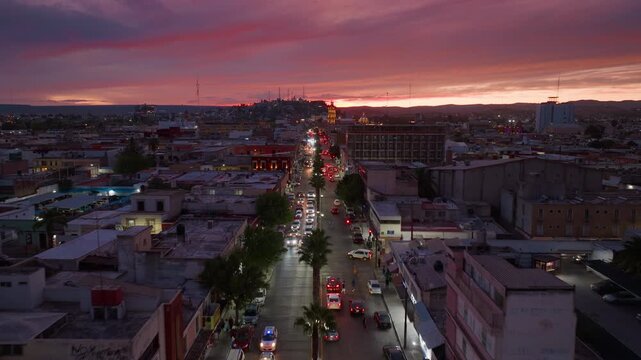 Aerial view of Durango Mexico downtown street at sunset with vibrant pink and orange sky. Cars drive along palm tree lined avenue with city buildings and rooftops visible in evening twilight.
