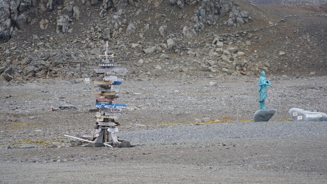 A wide shot of the iconic wooden distance signpost at the Russian Bellingshausen Station on King George Island, featuring a blue-patina bronze statue of Antarctic explorer Fabian von Bellingshausen