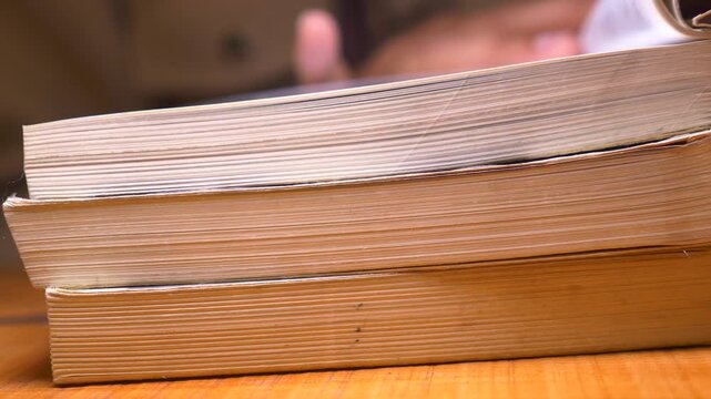 Stack of old books on table, person flipping through pages of magazine or book.