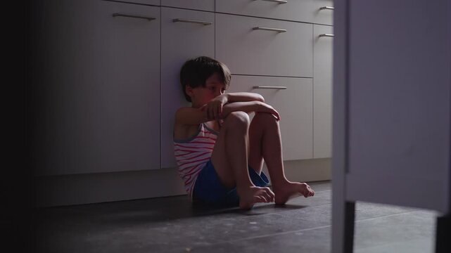 Young boy sitting against kitchen cabinet looking down at hands after crying, quiet childhood moment expressing sadness reflection vulnerability and emotional fatigue
