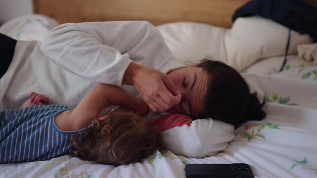 Mother lying on bed beside toddler child rubbing tired eye capturing moment of parental fatigue and exhaustion during quiet family rest time at home