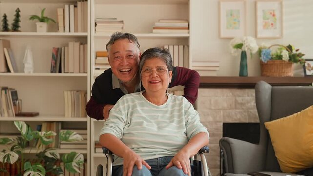 Portrait of elderly Asian husband supporting wife in wheelchair, both looking at camera with happiness, showing love, bonding, caregiving, strong relationship, family connection and warm retirement li