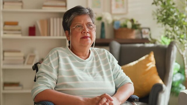 Elderly Asian woman in wheelchair looking out in living room, feeling lonely and missing freedom, retirement lifestyle change, emotional moment needing support, care, encouragement and family connecti