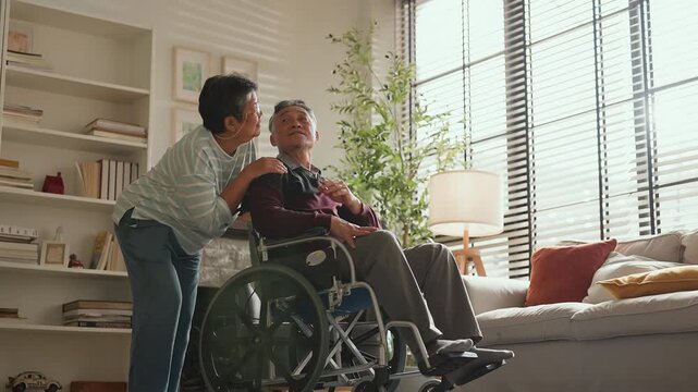Asian senior couple at home, retired man in wheelchair embraced by loving woman, caregiving support, bonding and true love, warm living room lifestyle, family connection, compassion and lifelong partn