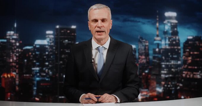 Authoritative Grey Haired Male Host in Dark Suit and Tie Sits at Desk, Looking at Camera and Gesturing Actively. Concept of Late Night Talk Show During Live Broadcast With Panoramic Night City View.