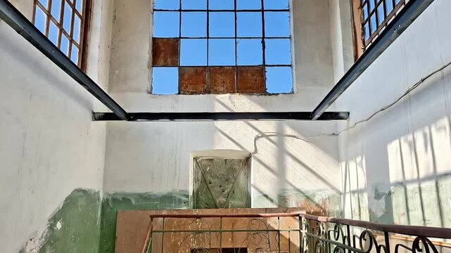 Sunlight pours through broken windows into an abandoned building, casting dramatic shadows on worn walls and railings, revealing textures of decay, silence, and forgotten urban history