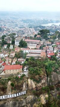 Aerial drone view above Antananarivo with the city skyline spread across surrounding hills, with dense urban neighborhoods and large historic palace complex overlooking Madagascar's capital. Vertical