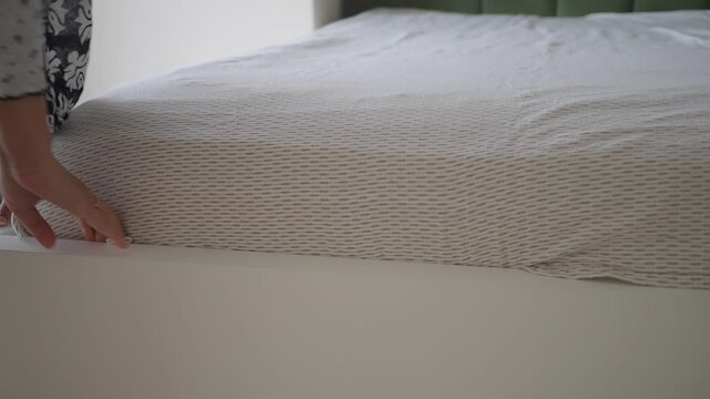 Woman hands tucking in grey patterned fitted sheet on white bed frame in bright bedroom area.
