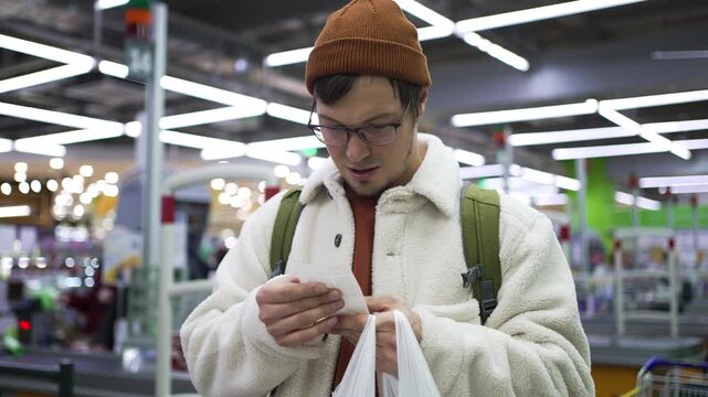 Worried young man in a supermarket feeling shocked and stressed while checking his expensive grocery receipt, showing concern over high food prices and inflation's impact on his budget