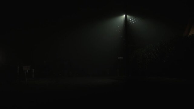 Foggy residential street at night with single street lamp creating dark cinematic atmosphere of quiet neighborhood with dense fog diffused light and low visibility
