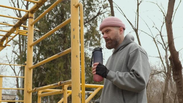 Mature man with beard taking a sip of water from sports bottle after intense training. Close up of male athlete in pink beanie and hoodie hydrating on outdoor fitness area