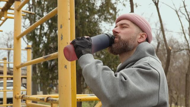Close up of bearded athlete drinking water from shaker bottle on sports ground. Focused man in pink beanie hydrating during morning workout session outdoors