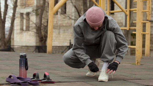 Man in pink beanie and grey tracksuit crouching to tie his shoelaces at outdoor fitness area. Male athlete preparing for street workout session on a cloudy day